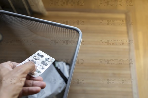 A hand discarding an pill blister pack into a mesh waste bin in a room with a wooden floor. The