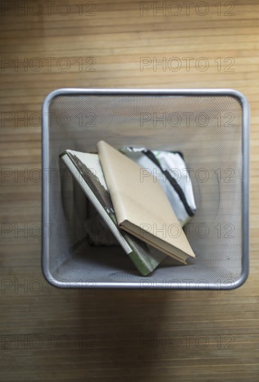 Aerial shot of a metal mesh waste bin containing discarded books, placed on a wooden floor. The