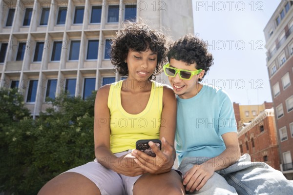 A multiethnic LGBTQ+ couple enjoys a sunny day in the city, looking at a smartphone together. The