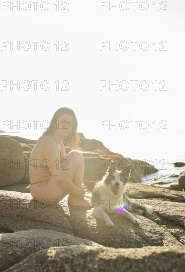A woman in a bikini sits beside her happy dog on sunlit rocks by the sea The peaceful setting