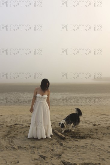 A woman in a white dress stands on a misty beach with a playful dog nearby The serene atmosphere