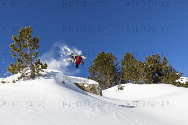 A snowboarder performs a backflip against a clear blue sky in a snow covered mountain landscape.