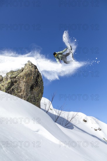 A snowboarder performs an aerial trick off a snowy cliff against a clear blue sky, capturing the