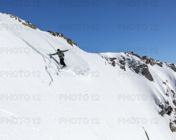 A snowboarder skillfully descends a steep, snowy slope against a clear blue sky. The untouched snow