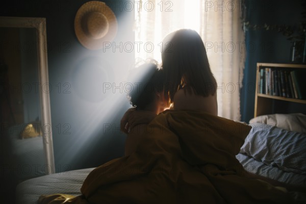 A couple shares an intimate embrace on a bed, bathed in warm morning light streaming through a