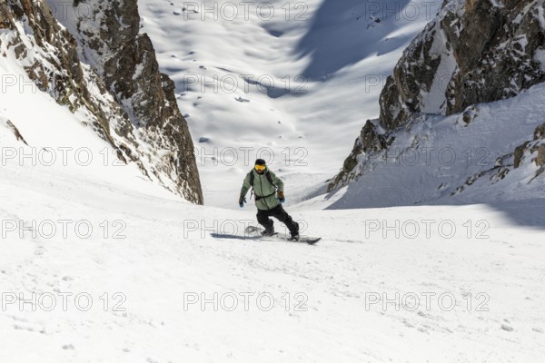 A snowboarder skillfully descends a steep, snowy mountain slope surrounded by rugged cliffs. The