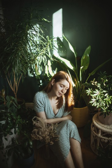 A woman in a light dress calmly sits surrounded by lush indoor plants. Sunlight filters through,