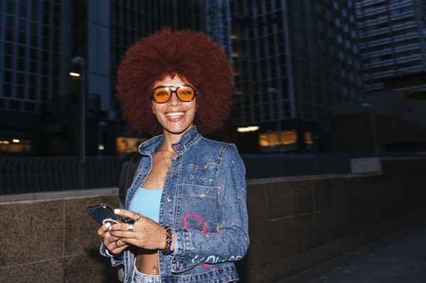 Fashionable African descendent woman with vibrant afro hair smiles while checking her smartphone in