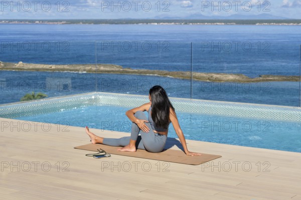A woman in athletic wear performs yoga on a mat by an infinity pool with a stunning ocean backdrop,