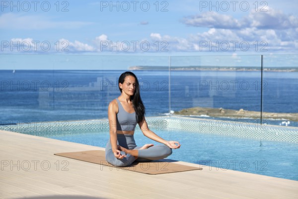 A woman practices yoga in lotus pose beside an infinity pool overlooking the ocean on a terrace.
