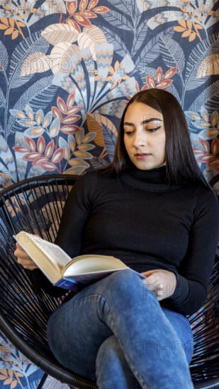 A young woman is engrossed in reading a book while sitting in a modern wicker chair, against a