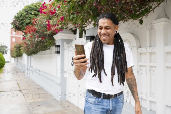 A joyful man with dreadlocks holds a smartphone, smiling widely as he stands in front of a vibrant,