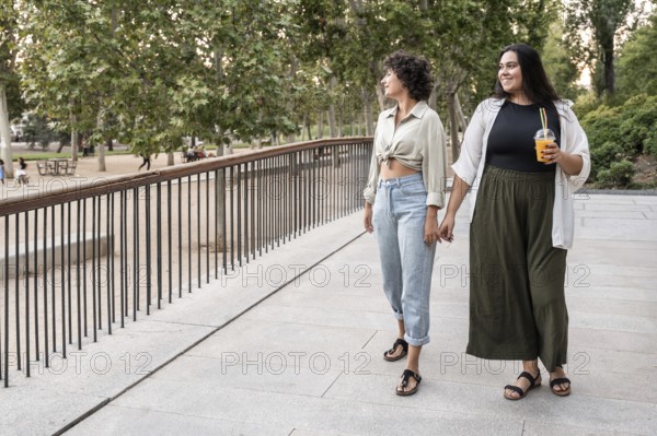 A lesbian couple walks hand in hand through a park, enjoying a leisurely stroll. One holds a drink,