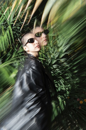 A stylish lesbian woman wearing sunglasses and a leather jacket stands amidst lush green foliage.