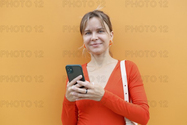 A woman stands against an orange wall in Warsaw, intently using her smartphone Dressed in a red top