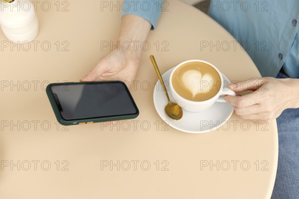 A woman relaxes in a Warsaw cafe, holding a smartphone and a cup of coffee with latte art The cozy