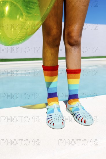 Vibrant scene featuring legs in rainbow socks and teal sandals, beside a pool. The bright turquoise