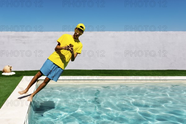 A teenage boy in vibrant yellow attire leans playfully by a pool, capturing a moment with a phone.