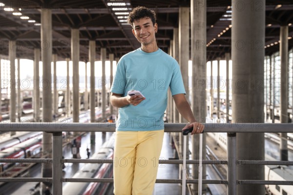 A young traveler in a vibrant blue shirt leans casually against a railing at a train station,