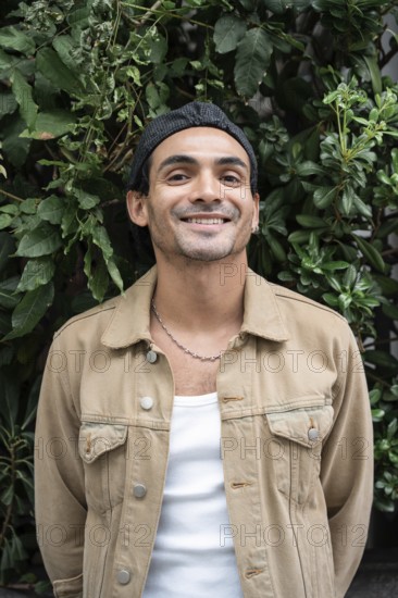 A gay man in casual clothing stands against a lush, leafy background. He sports a chain necklace