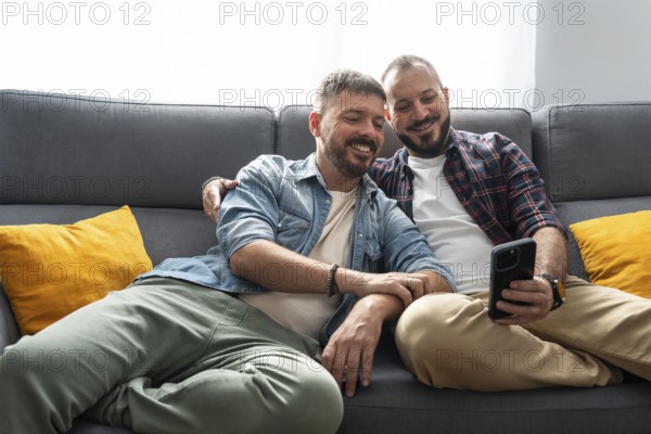 A joyful gay couple sits comfortably on the sofa, smiling as they look at a smartphone together in