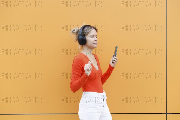 A young woman wearing headphones, joyfully listening to music and holding a smartphone She stands