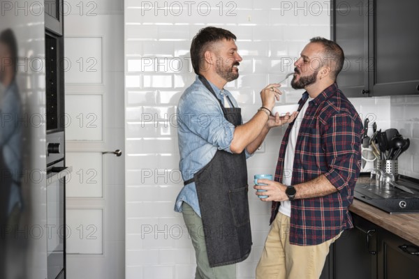 A joyful gay couple shares a moment in the kitchen, one feeding the other with a fork while smiling