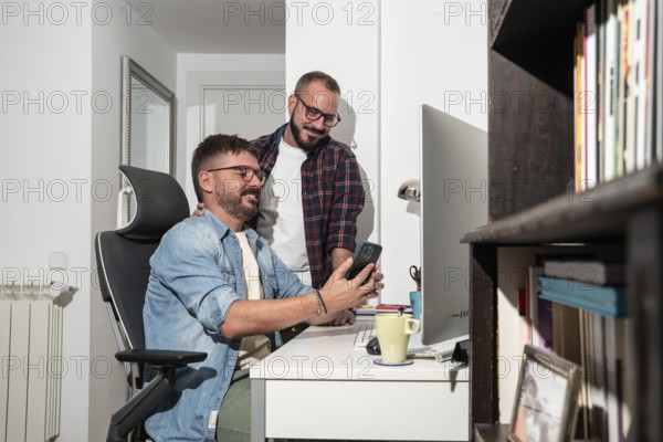 A joyful gay couple shares a moment at their home office. One man sits at a desk, holding a phone,