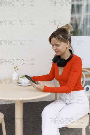 A woman sits at an outdoor cafe in Warsaw, using her smartphone and wearing headphones She enjoys a