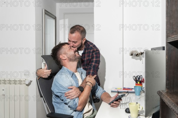 A joyful gay couple shares an affectionate moment in their home office. One partner sits with a