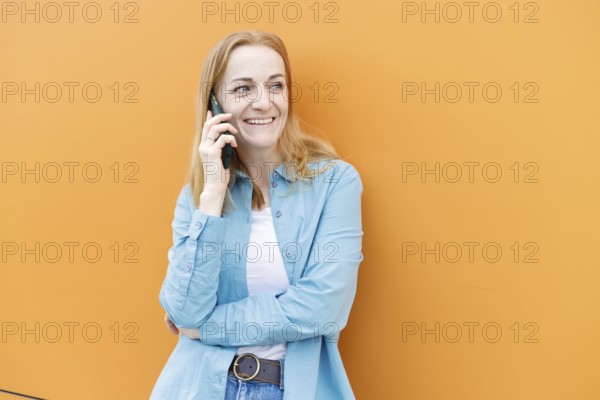 Smiling woman stands against a vibrant orange wall in Warsaw, chatting on a smartphone She wears