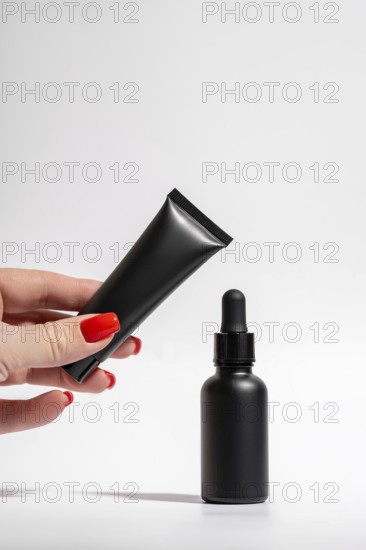 Cropped unrecognizable female hand with red manicure holding a matte black tube and dropper bottle
