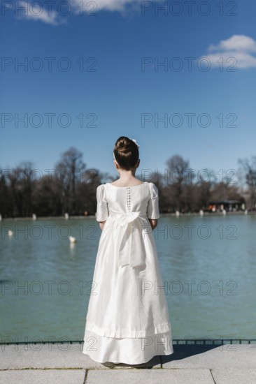 A girl stands by a serene lakeside, wearing a beautiful white First Communion Day dress. Her hair