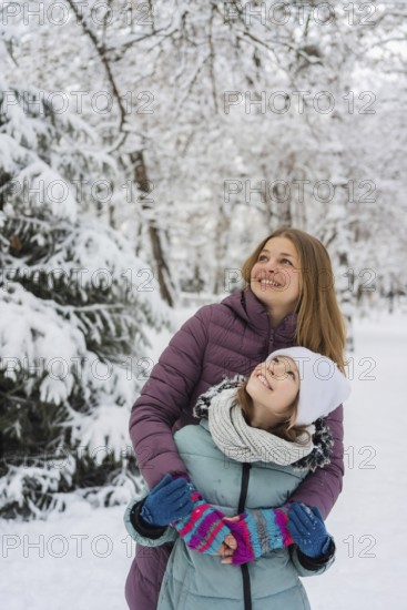A joyful mother embraces her daughter during a snowy winter day. They both wear colorful winter