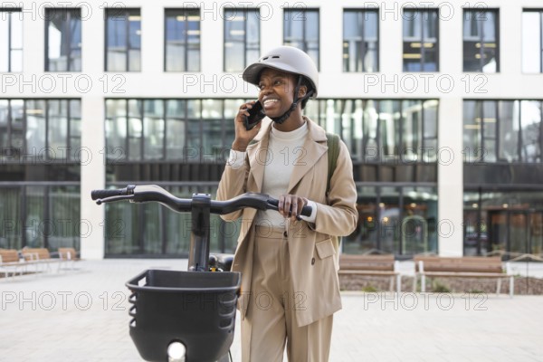 Smiling black woman in sleek beige suit and helmet, talking on a smartphone while holding a bike in