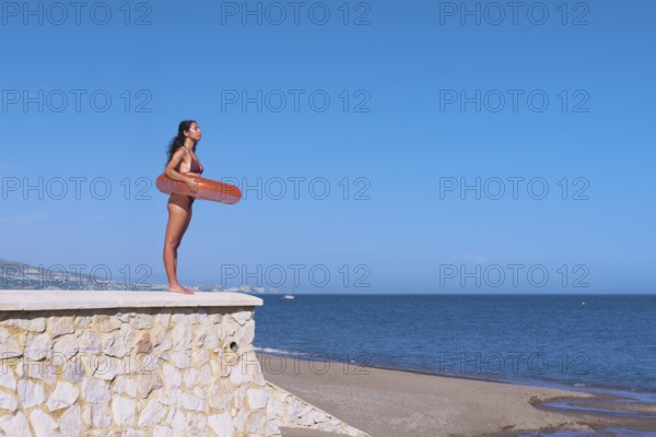 Side view of a young woman inside a float while standing atop a stone wall at the beach, gazing