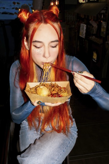 A woman with vibrant hair savoring noodles with chopsticks in an Asian food market, capturing the