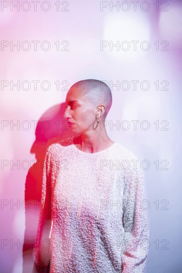 A middle-aged woman with a shaved head, wearing a textured white top, is illuminated by artistic