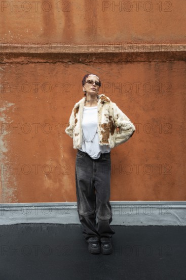 Stylish woman posing in a trendy fur jacket and sunglasses, set against a textured orange wall. The