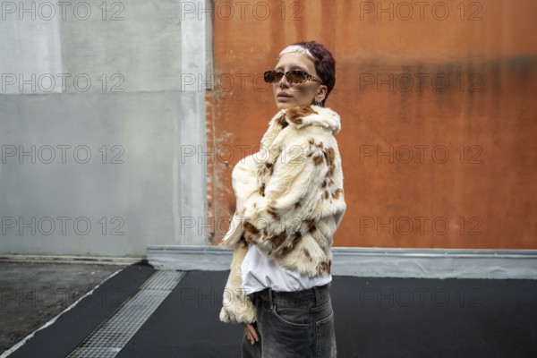 Fashionable woman in stylish faux fur jacket and sunglasses poses confidently against an urban