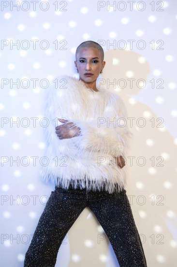 A middle-aged woman with a shaved head poses confidently. She wears a fur-like white jacket and