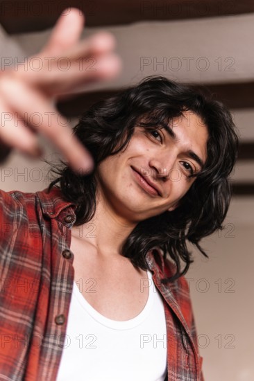A smiling young man in a white tank top and plaid shirt extends his hand towards the camera, giving