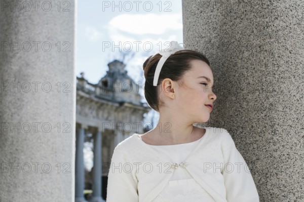 A young girl in a beautiful First Communion dress stands gracefully beside a large stone pillar,