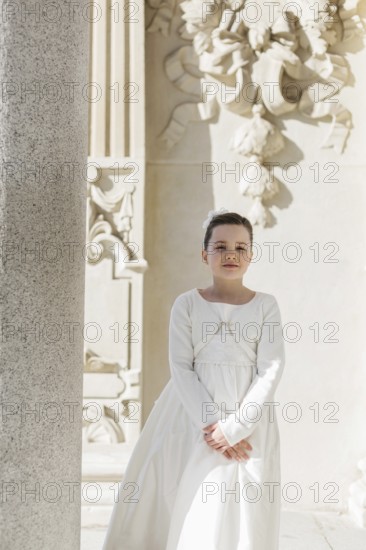 A young girl stands in her First Communion dress near an ornate architectural background, embodying
