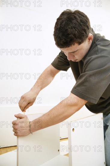 A focused young man assembles a new piece of white furniture using a screwdriver. He is seen
