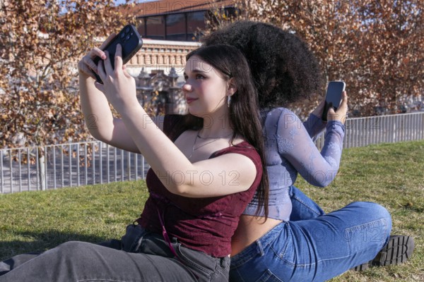 Two young women enjoy a sunny day outdoors while taking selfies. The lady on the left, with fair