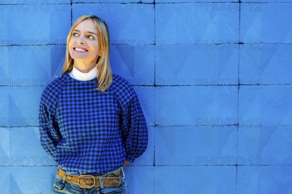 A cheerful woman smiles broadly while dressed in a blue checkered shirt and leaning against a