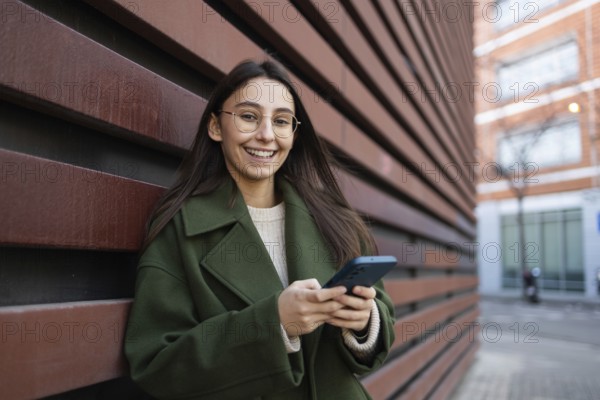 Young woman in a green coat smiling as she uses her smartphone, leaning against a red wall outdoors