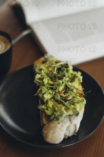 A close-up of avocado toast topped with greens and seeds, served on a black plate. A cup of coffee