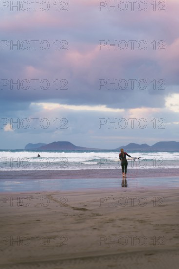 Surfers in a wetsuit approach the waves under a dramatic, colorful sky on a Lanzarote beach. The